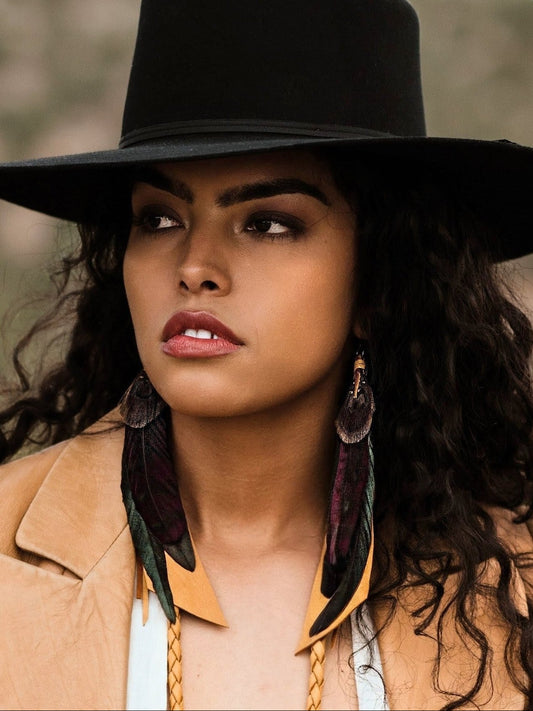 Woman wearing a black hat with feather earrings and a tan coat.